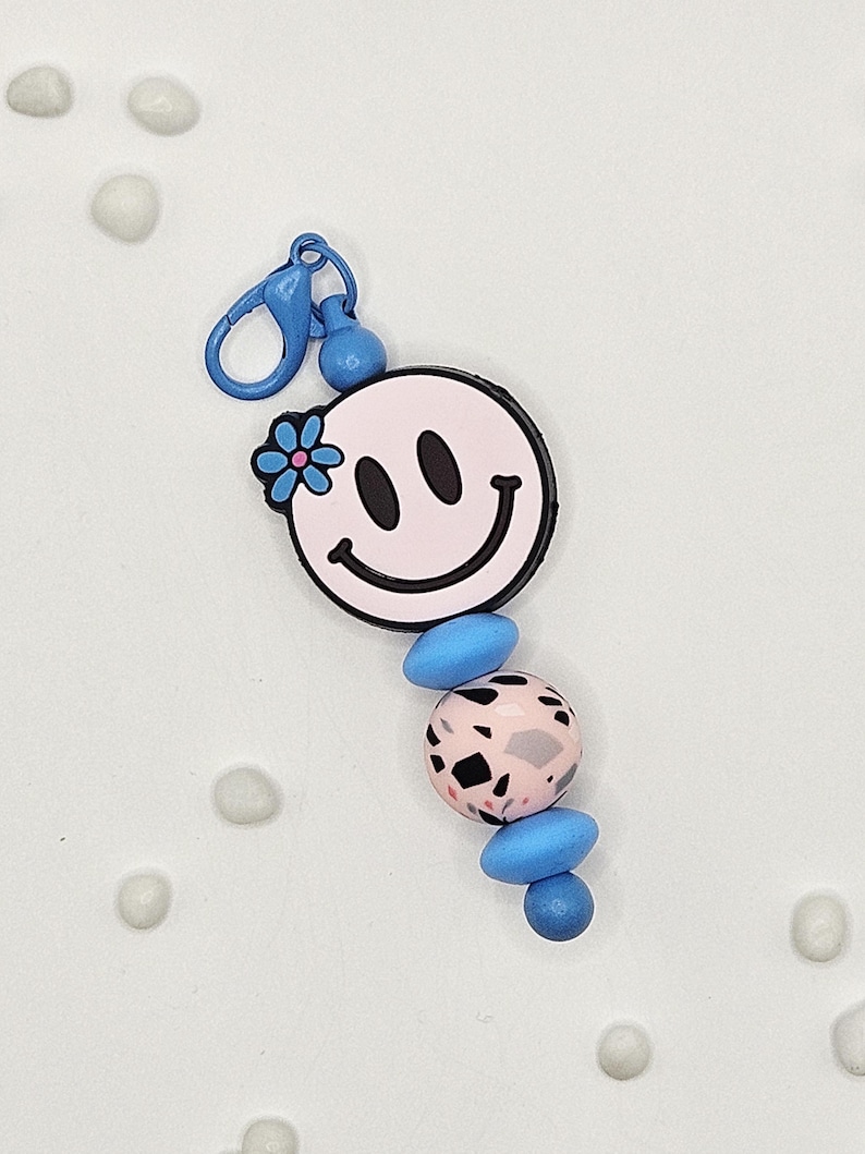 May include: A pink smiley face keychain with a blue flower and black outline. The keychain has a blue clasp and is adorned with blue and pink beads with black speckles. A fun and colorful accessory.