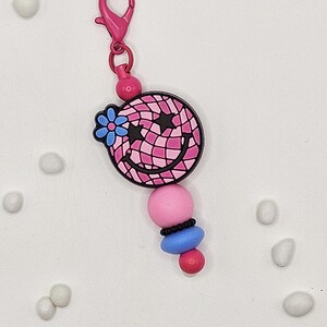 May include: A pink and black smiley face charm with a blue flower and star accents, attached to a pink lobster clasp. The charm has a pink, blue, and black bead design. A fun accessory for bags, keychains, or jewelry.