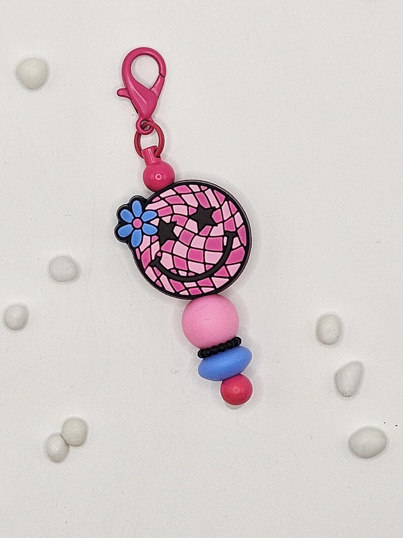 May include: A pink and black smiley face charm with a blue flower and star accents, attached to a pink lobster clasp. The charm has a pink, blue, and black bead design. A fun accessory for bags, keychains, or jewelry.