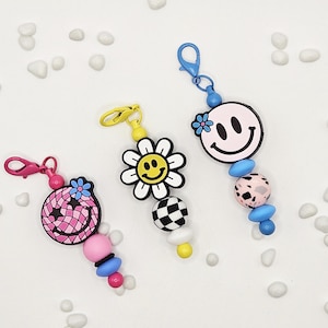 May include: Three colorful keychains with smiley face and flower designs. The keychains feature pink, yellow, and blue accents, with a variety of bead shapes and patterns. Each keychain has a clasp for easy attachment.