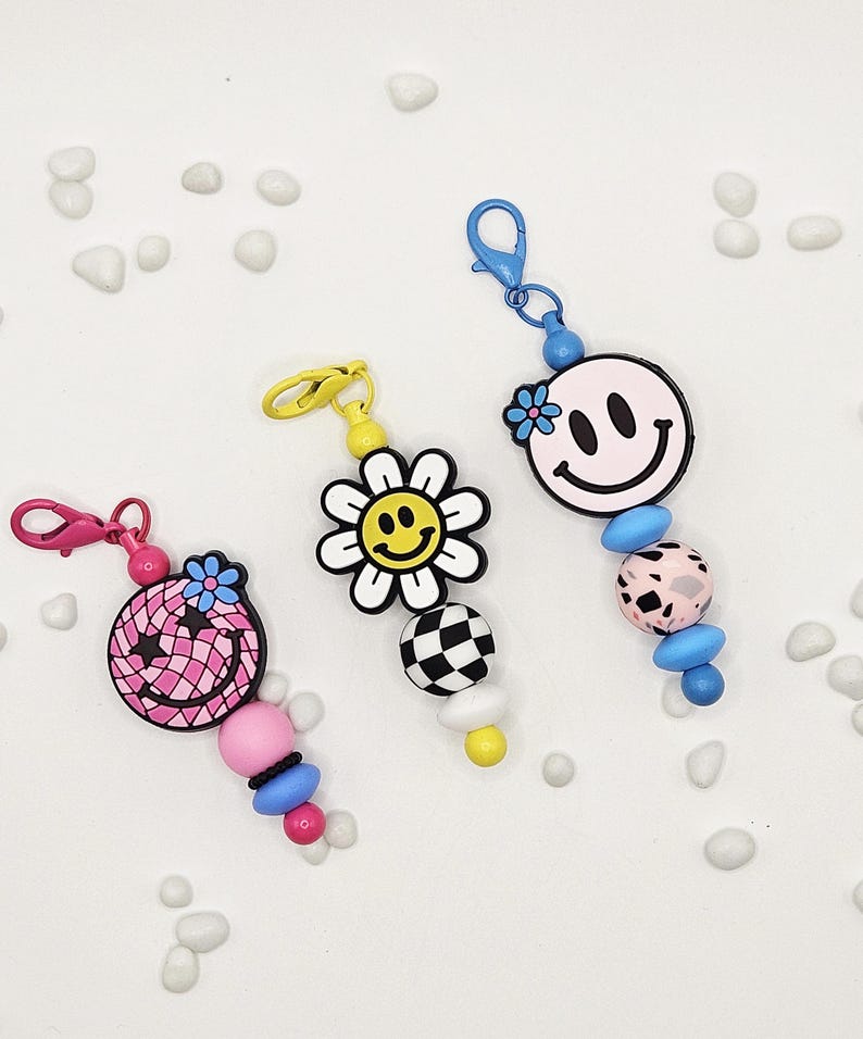 May include: Three colorful keychains with smiley face and flower designs. The keychains feature pink, yellow, and blue accents, with a variety of bead shapes and patterns. Each keychain has a clasp for easy attachment.