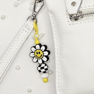 May include: A white leather crossbody bag with a silver zipper and a yellow and black daisy charm. The charm has a smiley face in the center of the daisy and a checkered bead below it. The bag has a white strap.
