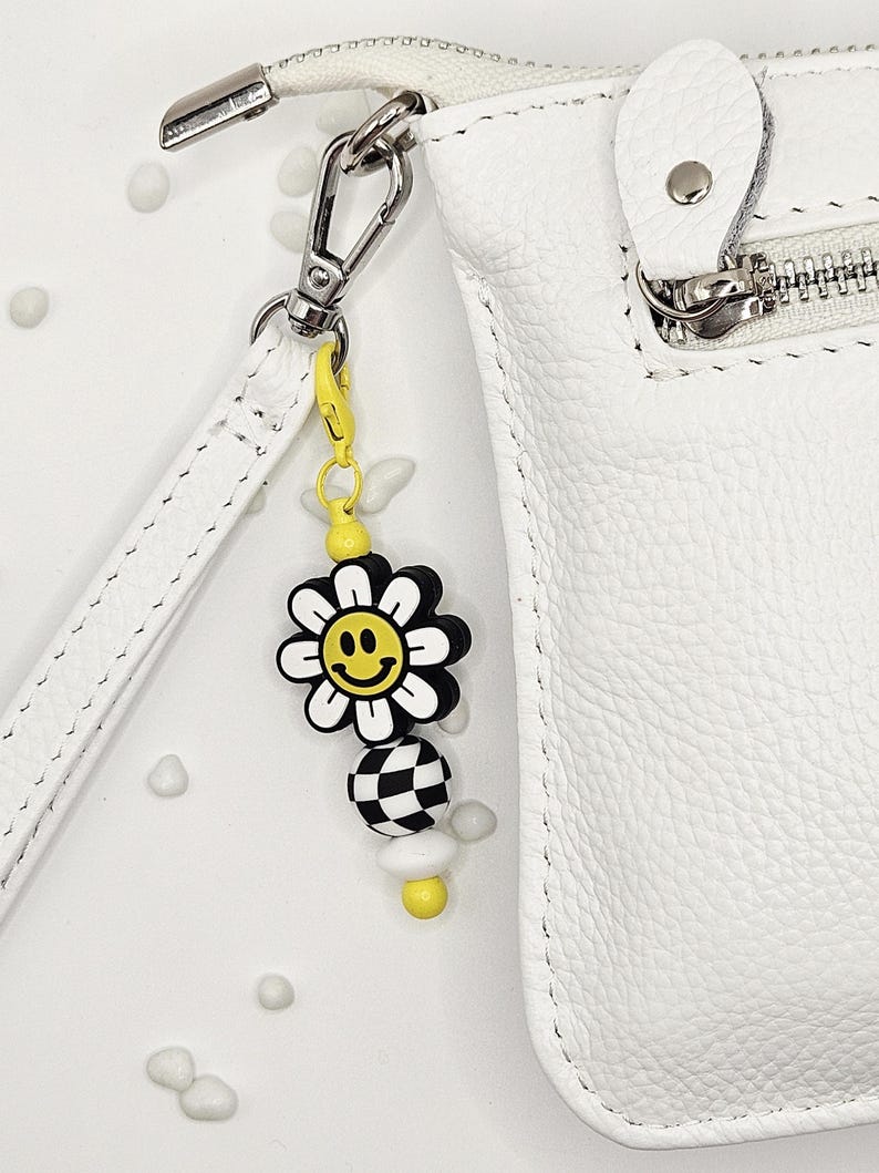 May include: A white leather crossbody bag with a silver zipper and a yellow and black daisy charm. The charm has a smiley face in the center of the daisy and a checkered bead below it. The bag has a white strap.