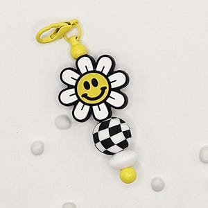 May include: A yellow and white daisy charm with a smiley face center, a black and white checkered ball, and a white mushroom-shaped bead. The charm has a yellow clasp and a yellow bead at the top.