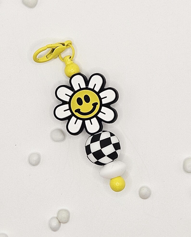 May include: A yellow and white daisy charm with a smiley face center, a black and white checkered ball, and a white mushroom-shaped bead. The charm has a yellow clasp and a yellow bead at the top.