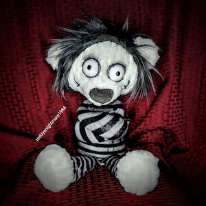 May include: A black and white striped plush toy with a stitched-on face and large eyes. The toy is sitting on a red fabric surface.