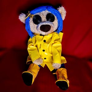 May include: A plush bear doll with blue yarn hair, button eyes, and a yellow raincoat. The bear wears matching yellow boots and a small gold hair clip. The background is a solid red color.