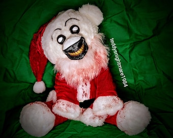 May include: A white teddy bear wearing a red Santa Claus suit with a white trim and a white beard. The bear has a creepy, painted-on smile and large, white eyes. The bear is lying on a green surface.