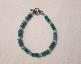 May include: Teal and light blue beaded bracelet with a silver-tone toggle clasp. The bracelet features alternating sections of teal cylindrical beads and light blue, translucent beads. The beads are strung together to form a flexible, wearable accessory.