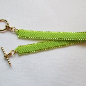 May include: A lime green beaded bracelet with gold-colored accents and a gold-tone toggle clasp. The bracelet is made of small, evenly spaced beads, creating a textured surface. The design features a loop on one end and a T-bar closure.
