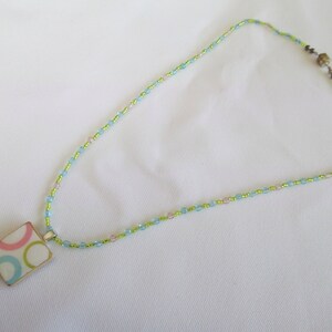 May include: A delicate necklace featuring a square pendant with a colorful circle pattern in pink, blue, and green. The pendant hangs from a beaded chain in shades of green, blue, and pink, creating a subtle, feminine accessory.