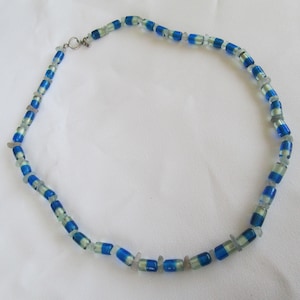 May include: A beaded necklace featuring a pattern of blue and light green cylindrical beads, interspersed with small, clear, irregular-shaped beads. The necklace has a silver-colored clasp. The jewelry is displayed on a white background.