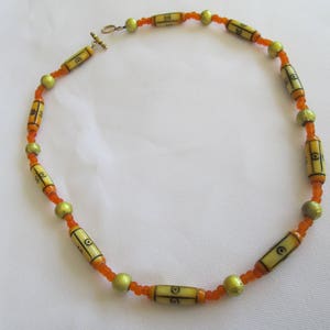 May include: A beaded necklace featuring alternating cylindrical beads with black geometric designs and small orange beads. The necklace also includes gold-colored spherical beads and a gold-tone clasp. The necklace is approximately 18 inches long.