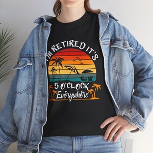 Puede incluir: Una camiseta negra con un gráfico de un atardecer en la playa con palmeras, una sombrilla y una persona en una silla. El texto I'M RETIRED IT'S 5 O'CLOCK Everywhere aparece en letras blancas y naranjas en la parte delantera.