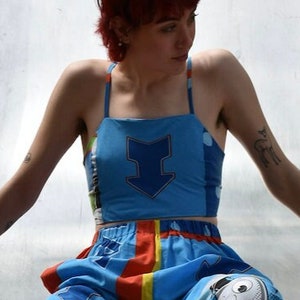 May include: A person wearing a blue and yellow patterned crop top with a blue arrow design and matching pants. The person is sitting on a metal slide.