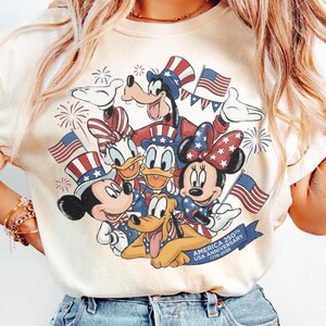 May include: Cream-coloured t-shirt with a graphic of Disney characters Mickey Mouse, Minnie Mouse, Donald Duck, Daisy Duck, Goofy, and Pluto, in patriotic outfits. The design includes American flags and fireworks, with text reading "America 250th USA Anniversary 1776-2026".