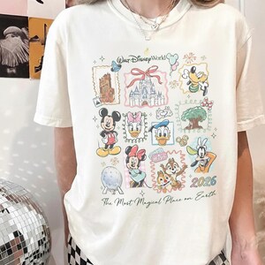 May include: Cream-colored t-shirt featuring a colorful collage of Disney characters and landmarks. The design includes Mickey Mouse, Minnie Mouse, Donald Duck, and Goofy, with the text "Walt Disney World" and "2026". The shirt also has the phrase "The Most Magical Place on Earth".