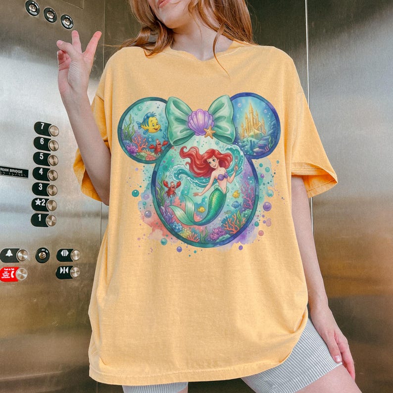 May include: Yellow t-shirt featuring a colorful graphic of a mermaid inside a Mickey Mouse head shape. The design includes a mermaid, a castle, and sea creatures. The shirt has short sleeves and a relaxed fit.