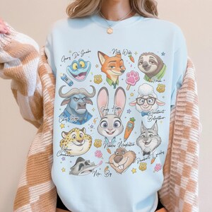 K&ouml;nnte beinhalten: Ein hellblaues Sweatshirt mit Rundhalsausschnitt und einem Grafikdruck verschiedener animierter Tierfiguren, darunter ein Hase, ein Fuchs, ein Faultier und ein Leopard. Das Design enth&auml;lt Textbeschriftungen, kleine Polizeimarken, Karotten und Pfotenabdr&uuml;cke.