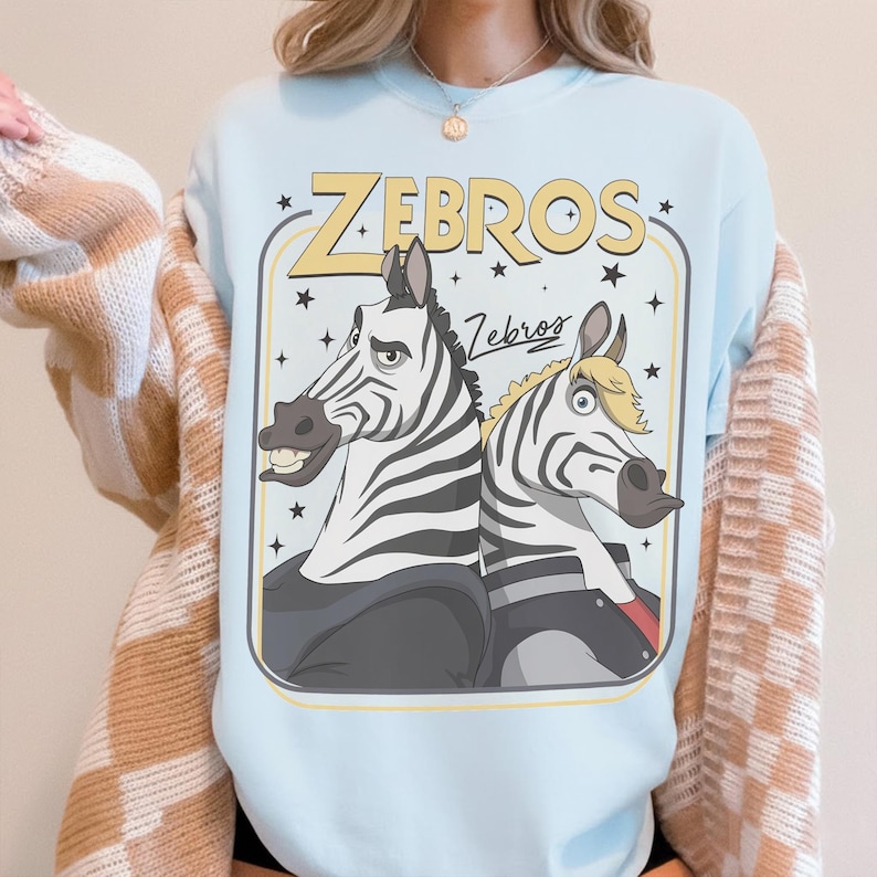 K&ouml;nnte beinhalten: Ein hellblaues Sweatshirt mit Rundhalsausschnitt und einem Grafikdruck von zwei Zebras im Cartoon-Stil, die R&uuml;cken an R&uuml;cken stehen. Der Text "ZEBROS" steht in gro&szlig;en gelben Buchstaben dar&uuml;ber, erg&auml;nzt durch das Wort "Zebros" in Schreibschrift.