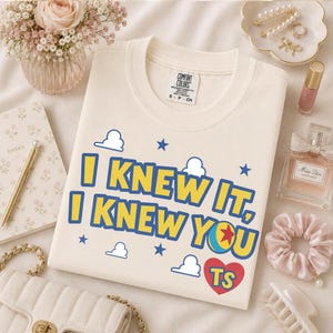 Pu&ograve; includere: Una t-shirt a maniche corte color crema con il testo blu e giallo "I KNEW IT, I KNEW YOU", un cuore rosso contenente le lettere "TS" e una piccola grafica a forma di palla. La maglietta include un'etichetta del marchio Comfort Colors taglia S.