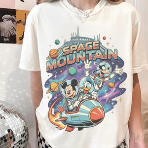 Może przedstawiać: Kremowa koszulka z kolorową grafiką Myszki Miki, Kaczora Donalda i Goofy'ego w skafandrach kosmicznych jadących rakietą przed Space Mountain. Napis "SPACE MOUNTAIN" jest wyświetlany nad postaciami w stylu retro.