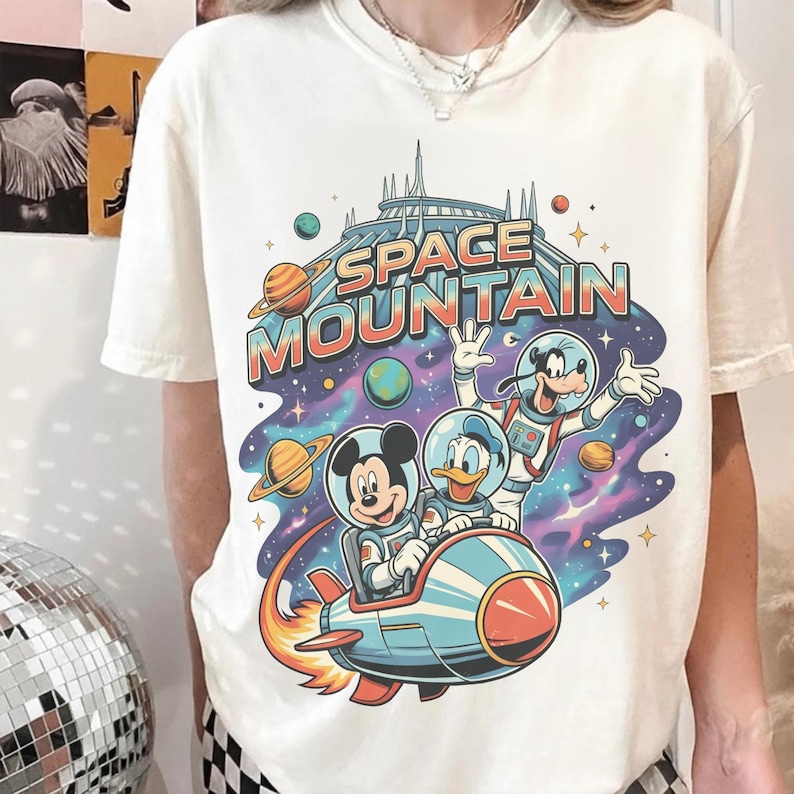 Może przedstawiać: Kremowa koszulka z kolorową grafiką Myszki Miki, Kaczora Donalda i Goofy'ego w skafandrach kosmicznych jadących rakietą przed Space Mountain. Napis "SPACE MOUNTAIN" jest wyświetlany nad postaciami w stylu retro.