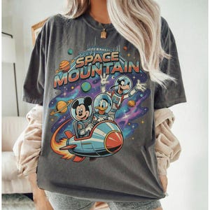 Może przedstawiać: Ciemnoszara koszulka z kolorową grafiką Myszki Miki, Kaczora Donalda i Goofy'ego w skafandrach kosmicznych na rakiecie, z napisem "SPACE MOUNTAIN". Projekt zawiera planety i gwiazdy.