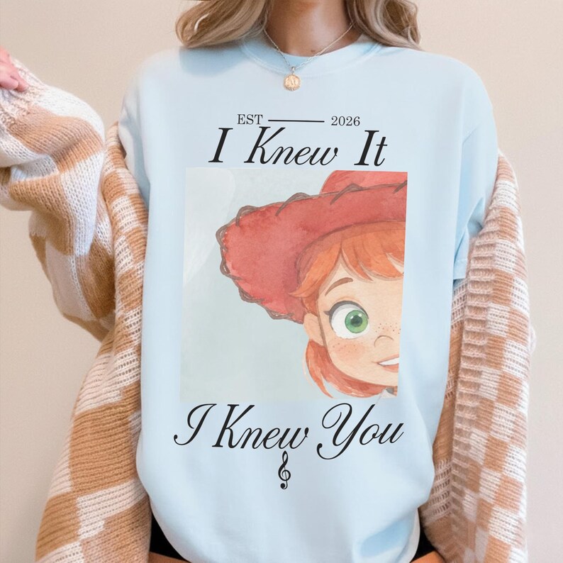 May include: A light blue crewneck sweatshirt featuring a graphic print of a cartoon character with red hair and a red hat. The text reads EST 2026 I Knew It I Knew You with a musical treble clef symbol below the text. The shirt is worn under a tan cardigan.