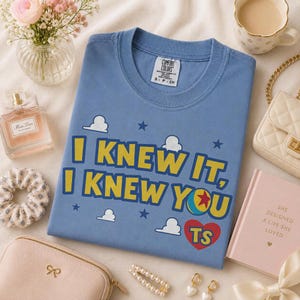 Pu&ograve; includere: Una t-shirt blu a girocollo con il testo giallo I KNEW IT, I KNEW YOU, un'icona a forma di palla colorata, un cuore rosso con le lettere TS e grafiche di nuvole bianche e stelle blu. L'etichetta Comfort Colors &egrave; presente sul colletto.