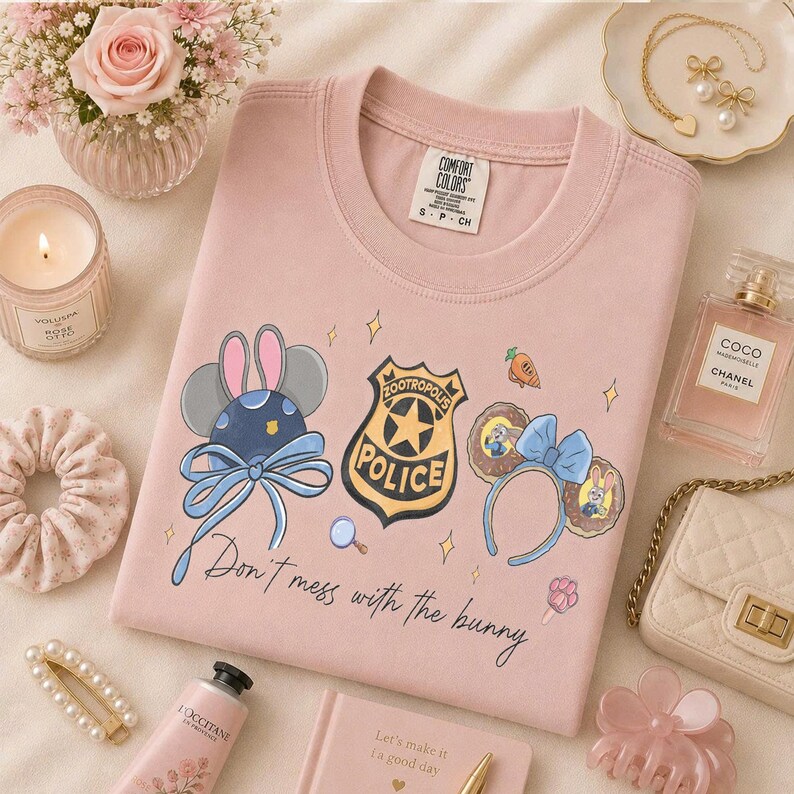 May include: A light pink t-shirt featuring a Zootropolis Police badge, two sets of mouse ears, and the text "Don't mess with the bunny" in black script. The shirt has a crew neckline and a Comfort Colors brand label on the inner collar.