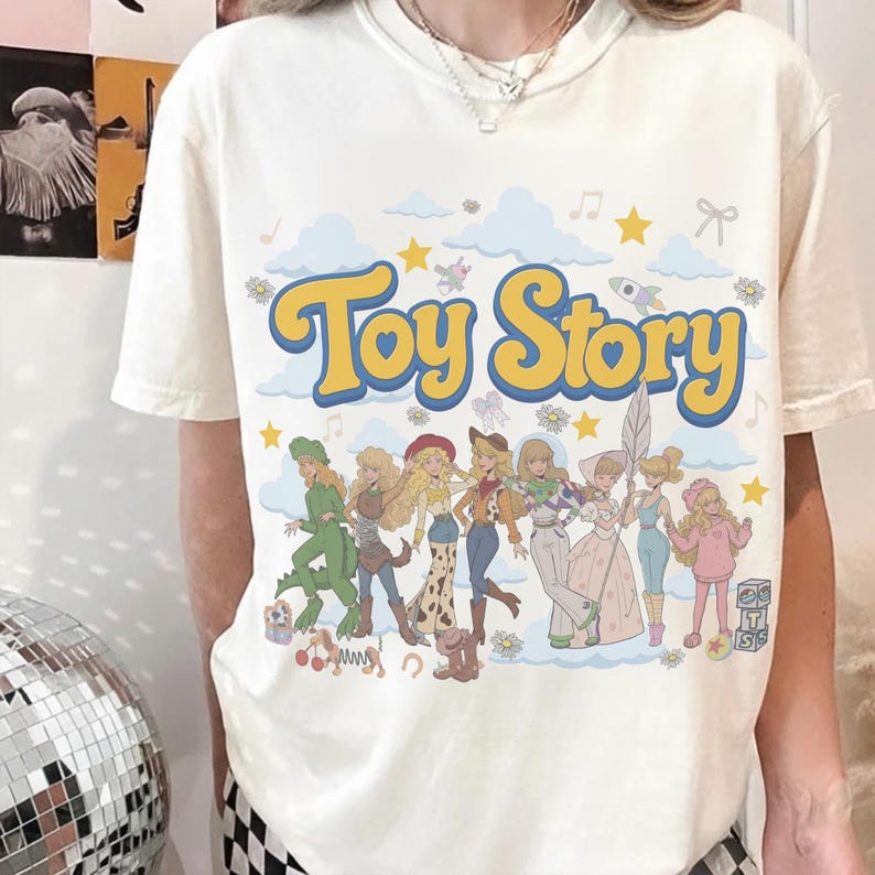 May include: A cream-coloured short-sleeve t-shirt featuring the text Toy Story in large yellow letters with blue outlines. Below the text is a graphic illustration of eight characters in a stylised aesthetic, surrounded by clouds, stars, and flowers.