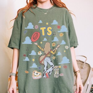 May include: An olive green t-shirt featuring a graphic print of a person with long hair playing a guitar, wearing a yellow plaid shirt and cow-print trousers. The design includes the text TS, a red hat, blue clouds, flowers, stars, and blocks.