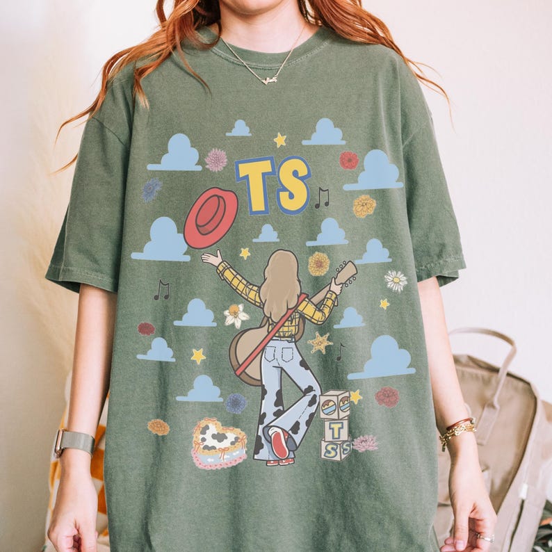 May include: An olive green t-shirt featuring a graphic print of a person with long hair playing a guitar, wearing a yellow plaid shirt and cow-print trousers. The design includes the text TS, a red hat, blue clouds, flowers, stars, and blocks.