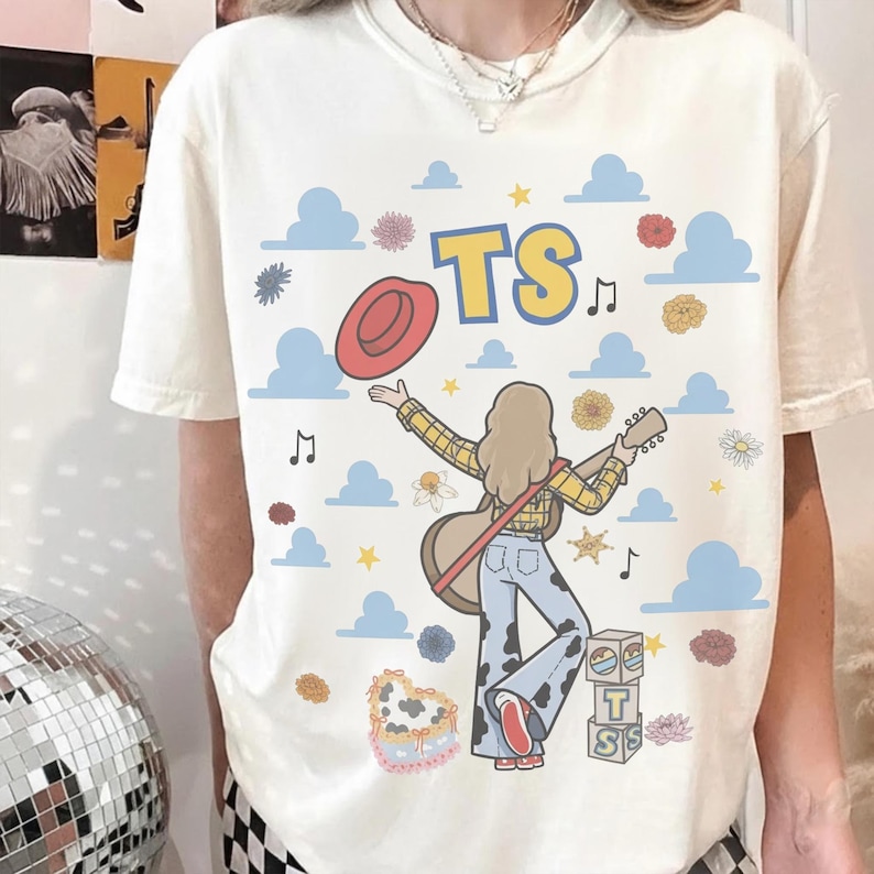 May include: A cream-coloured t-shirt featuring a graphic of a person with long hair wearing a yellow plaid shirt and cow-print bell-bottom jeans, holding a guitar and a red hat. The design includes blue clouds, flowers, musical notes, and the text TS.