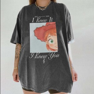 May include: A charcoal gray short-sleeve t-shirt featuring a graphic print of a cartoon character with red hair and a red hat. The text reads "EST 2026 I Knew It I Knew You" with a small musical treble clef symbol centered below the text.