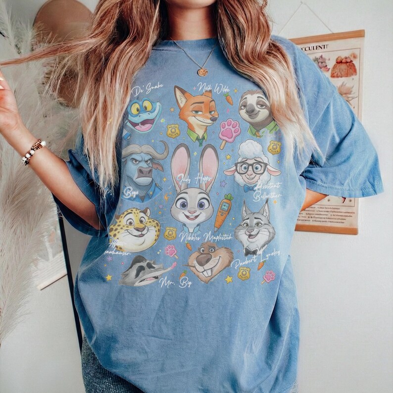 K&ouml;nnte beinhalten: Ein blaues Kurzarm-T-Shirt mit einem Grafikdruck verschiedener animierter Tierfiguren, darunter ein Hase, ein Fuchs und ein Faultier. Das Design enth&auml;lt Textbeschriftungen f&uuml;r jede Figur sowie kleine Symbole von Karotten, Pfoten und Abzeichen.