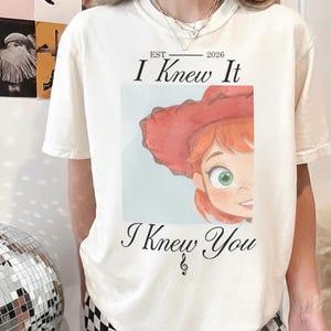 May include: An off-white short-sleeve t-shirt featuring a graphic of a red-haired character wearing a red cowboy hat. The text reads EST 2026, I Knew It, and I Knew You, with a small musical note symbol below the text on the front of the shirt.