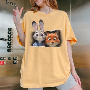 K&ouml;nnte beinhalten: Senfgelbes T-Shirt mit einem Aufdruck von Judy Hopps und Nick Wilde aus Zoomania. Die Zeichentrickfiguren sind dargestellt, wie sie aus einem zerrissenen Papiereffekt auftauchen. Das Shirt hat kurze &Auml;rmel und eine lockere Passform.