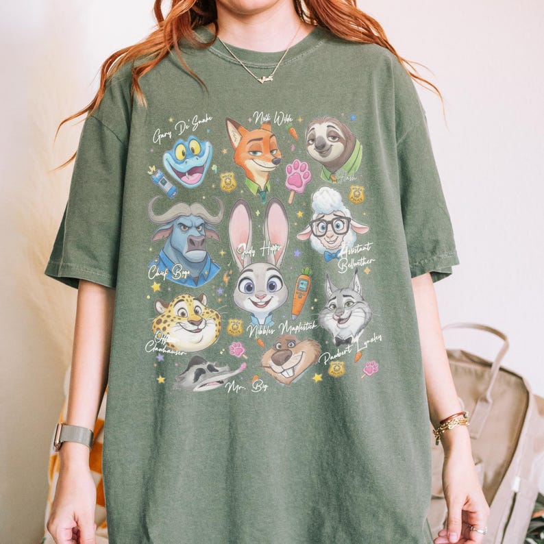 K&ouml;nnte beinhalten: Ein olivgr&uuml;nes Kurzarm-T-Shirt mit einem Grafikdruck verschiedener animierter Tierfiguren aus Zoomania, darunter Judy Hopps, Nick Wilde und Chief Bogo, erg&auml;nzt durch kleine Symbole wie einen Karottenstift und einen Pfotenabdruck.