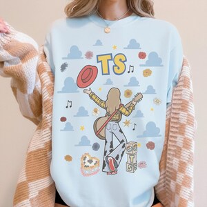 May include: A light blue crewneck sweatshirt featuring a graphic illustration of a person with long hair, wearing a yellow plaid shirt and cow-print trousers, holding a guitar and a red hat. The design includes blue clouds, flowers, and the text TS.
