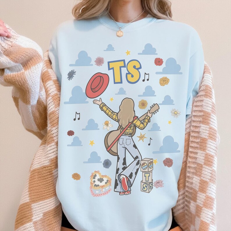 May include: A light blue crewneck sweatshirt featuring a graphic illustration of a person with long hair, wearing a yellow plaid shirt and cow-print trousers, holding a guitar and a red hat. The design includes blue clouds, flowers, and the text TS.