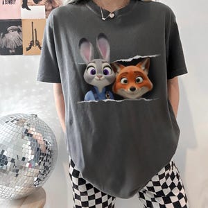 K&ouml;nnte beinhalten: Dunkelgraues T-Shirt mit einem Aufdruck von Judy Hopps und Nick Wilde aus Zoomania. Die Zeichentrickfiguren scheinen aus einem zerrissenen Bereich herauszukommen. Das T-Shirt hat kurze &Auml;rmel und eine lockere Passform.
