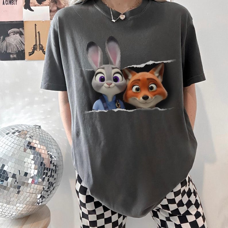 K&ouml;nnte beinhalten: Dunkelgraues T-Shirt mit einem Aufdruck von Judy Hopps und Nick Wilde aus Zoomania. Die Zeichentrickfiguren scheinen aus einem zerrissenen Bereich herauszukommen. Das T-Shirt hat kurze &Auml;rmel und eine lockere Passform.
