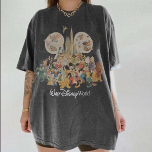 May include: Dark gray t-shirt featuring a colorful graphic of Disney characters and Cinderella's castle. The text "Walt Disney World" is printed below the graphic. The shirt has short sleeves and a relaxed fit.