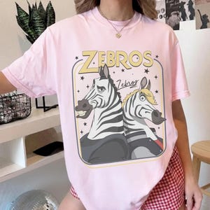 K&ouml;nnte beinhalten: Ein hellrosa T-Shirt mit kurzen &Auml;rmeln und einem Grafikdruck von zwei Zeichentrickzebras in einem rechteckigen Rahmen. Der Text "ZEBROS" steht in gro&szlig;en gelben Buchstaben &uuml;ber den Zebras, darunter "Zebros" in Schreibschrift. Die Zebras haben schwarze und wei&szlig;e Streifen.