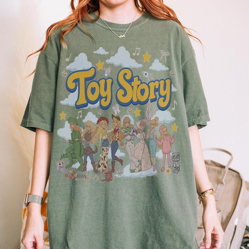 May include: An olive green short-sleeve t-shirt featuring a large graphic print with the text "Toy Story" in yellow letters. The design includes a group of characters dressed in various costumes, surrounded by clouds, stars, and musical notes.
