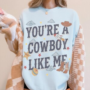 May include: A light blue crewneck sweatshirt featuring the dark blue text "YOU'RE A COWBOY LIKE ME" surrounded by graphic illustrations of a brown cowboy hat, a rocket ship, a yellow sheriff star, a ball, and brown cowboy boots with clouds and stars.