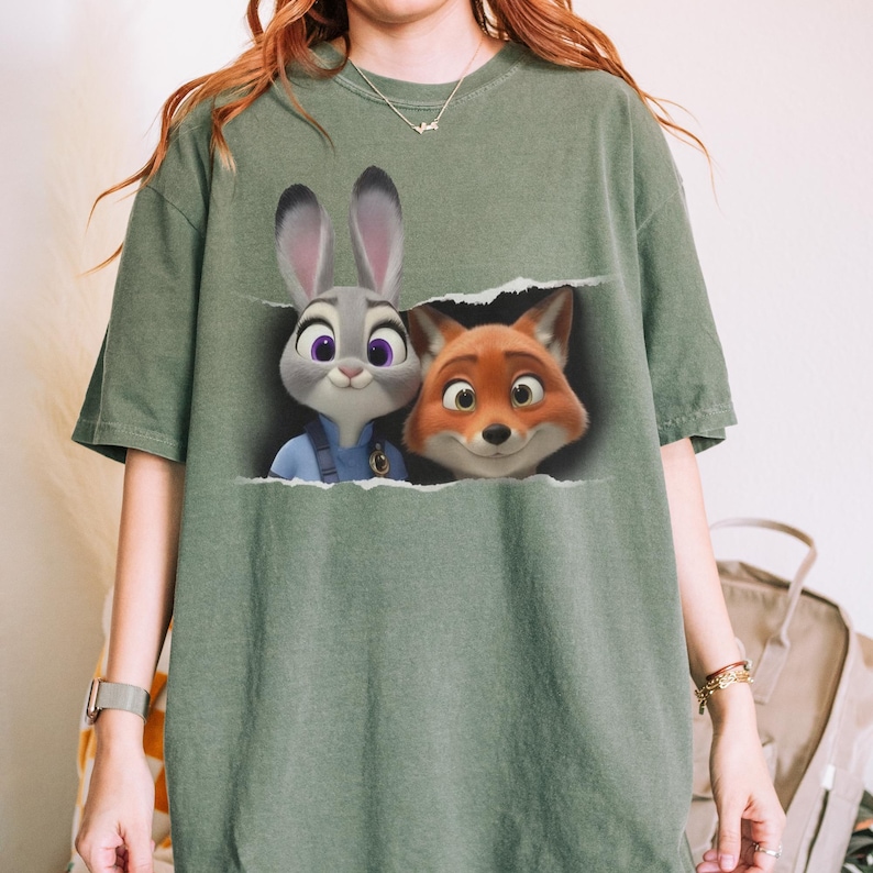 K&ouml;nnte beinhalten: Ein olivgr&uuml;nes T-Shirt mit einer Grafik von Judy Hopps und Nick Wilde aus Zoomania. Die Charaktere sind zu sehen, wie sie durch einen zerrissenen Teil des Shirts blicken. Das Shirt hat kurze &Auml;rmel und eine lockere Passform.