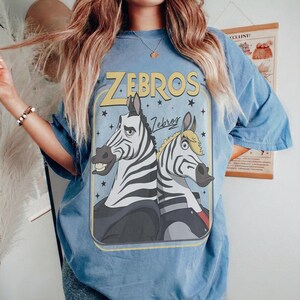 K&ouml;nnte beinhalten: Ein blaues T-Shirt mit einem Grafikdruck von zwei Zebras und dem Text "ZEBROS" sowie "Zebros" in Gelb und Schwarz. Die Zebras sind im Cartoon-Stil dargestellt, eines mit einer dunklen Jacke und das andere mit blondem Haar, vor einem Hintergrund aus Sternen.